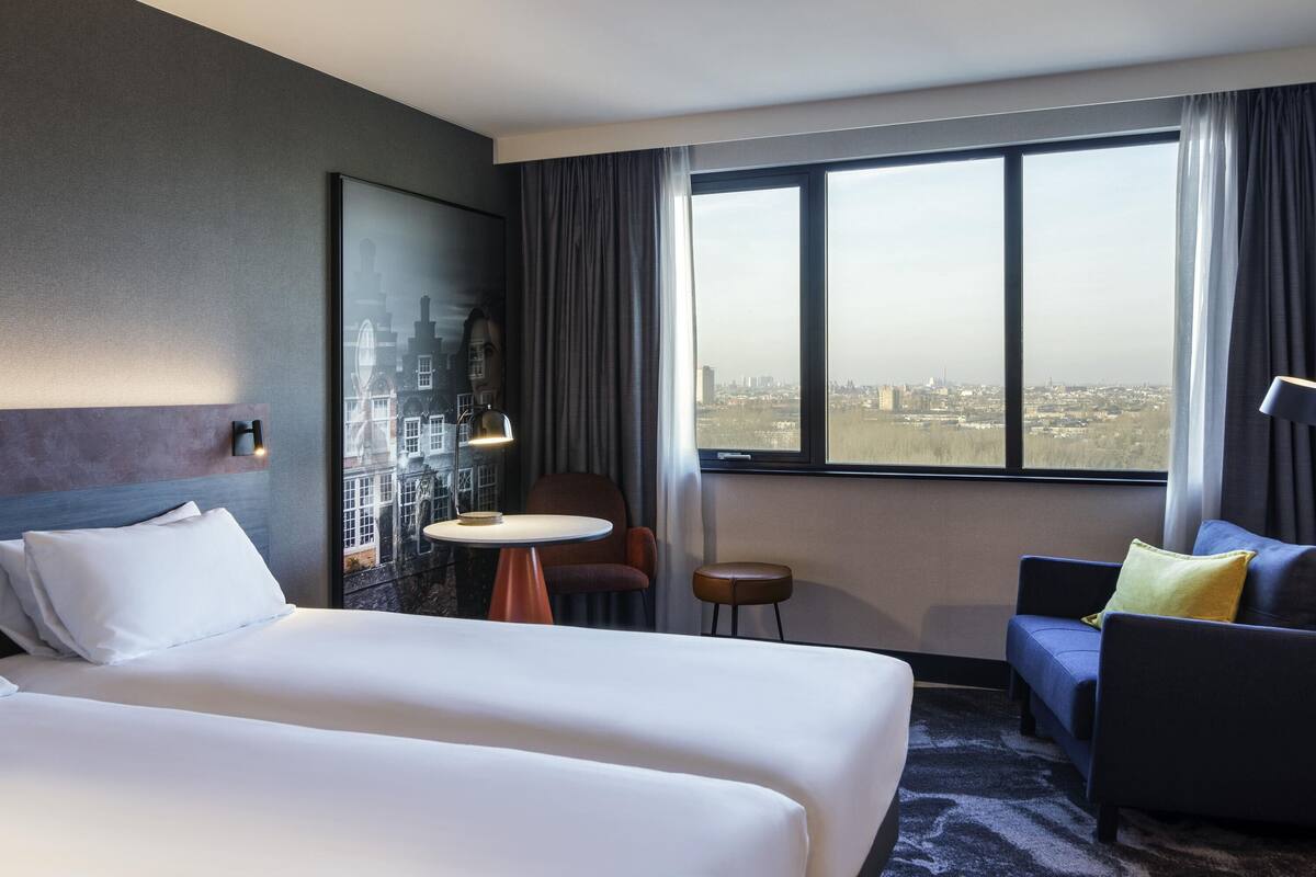 Habitación de hotel con cama, una mesita, un sillón, un sofá y un amplio ventanal con vistas a la ciudad.
