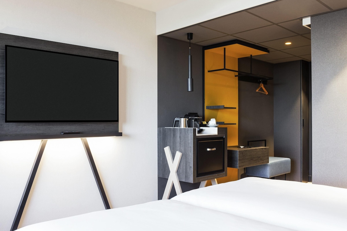 Una habitación de hotel moderna con una amplia televisión de pantalla plana montada en la pared, una cama con ropa blanca, un escritorio pequeño con cafetera y un espejo sobre el lavabo.