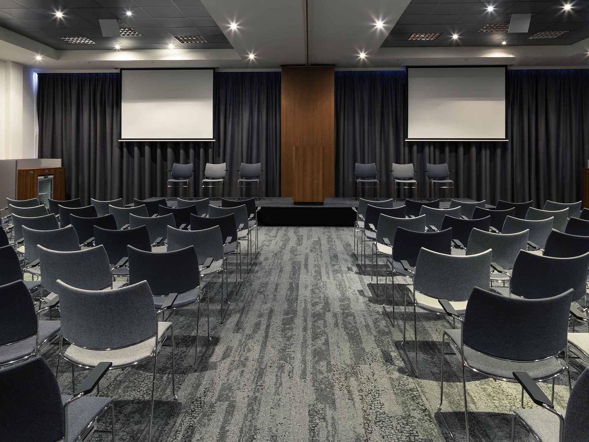 Een congrescentrum met rijen stoelen, een spreekgestoelte en twee schermen aan de achterwand.