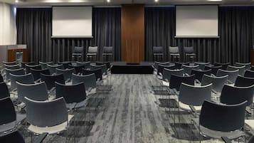 Een congrescentrum met rijen stoelen, een spreekgestoelte en twee schermen aan de achterwand.