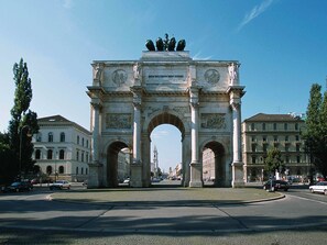 Un gran arco de piedra con inscripciones y estatuas, rodeado de edificios y una calle con coches.