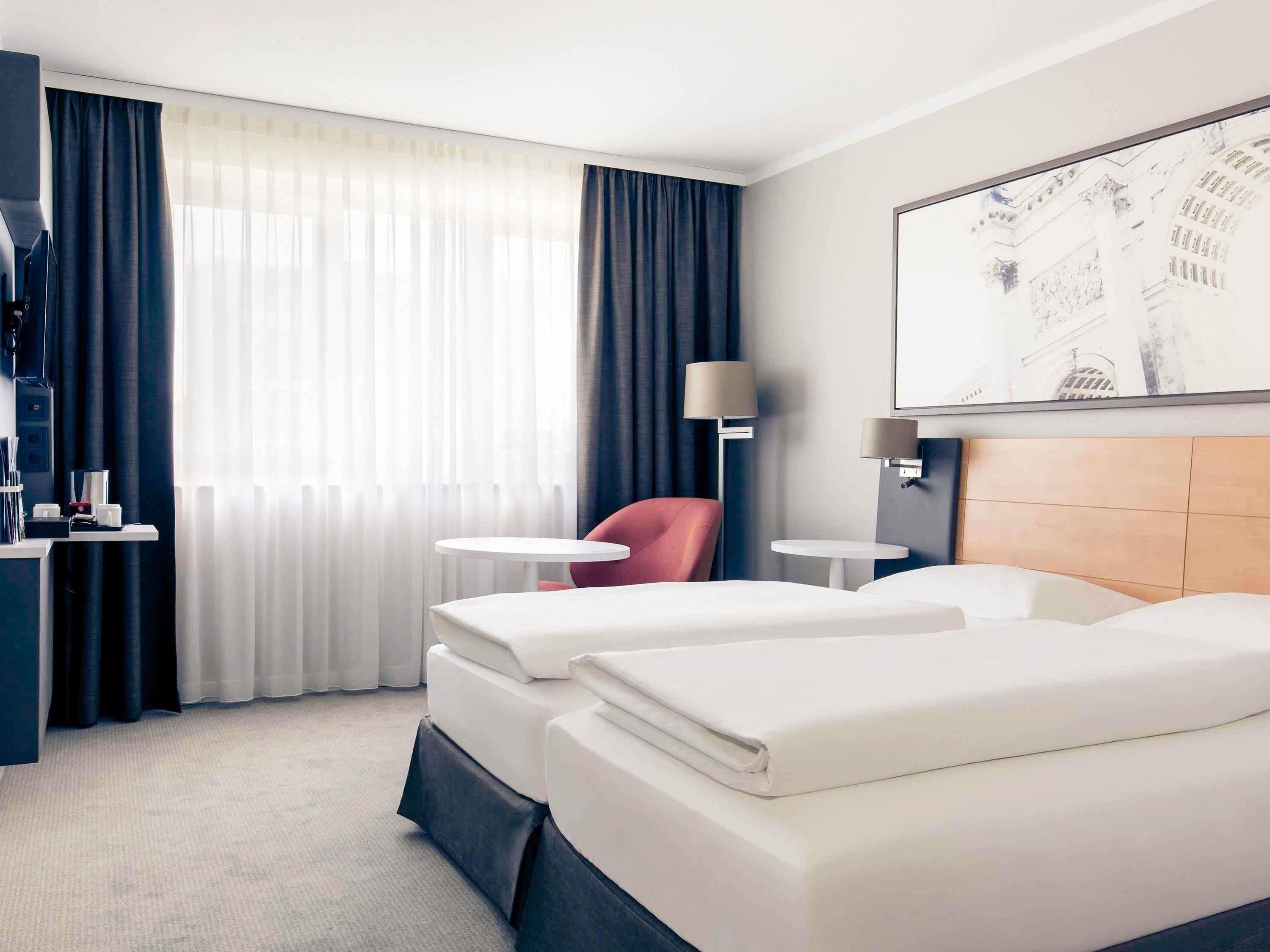 Habitación de hotel con dos camas, un escritorio con lámpara, una silla y un amplio ventanal con cortinas.
