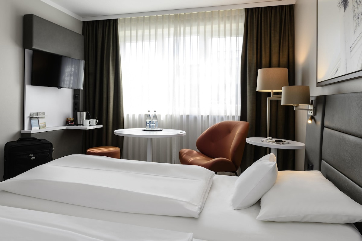 A hotel room with a bed, a desk, a chair, a round table, a TV, and a window with curtains.