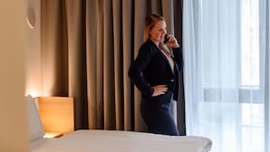 A hotel room with a bed, a nightstand, a lamp, and a window with curtains. - Mercure München City Schwabing (Munich)