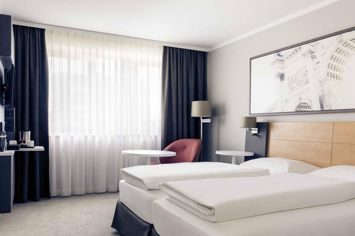Een hotelkamer met twee bedden, een bureau met lamp, een stoel en een groot raam met gordijnen.