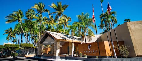 A hotel entrance with palm trees, a covered waiting area, and flags.