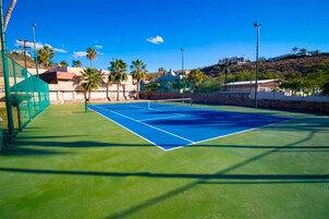 Una cancha de tenis con una red, rodeada por una cerca verde, palmeras y casas al fondo.