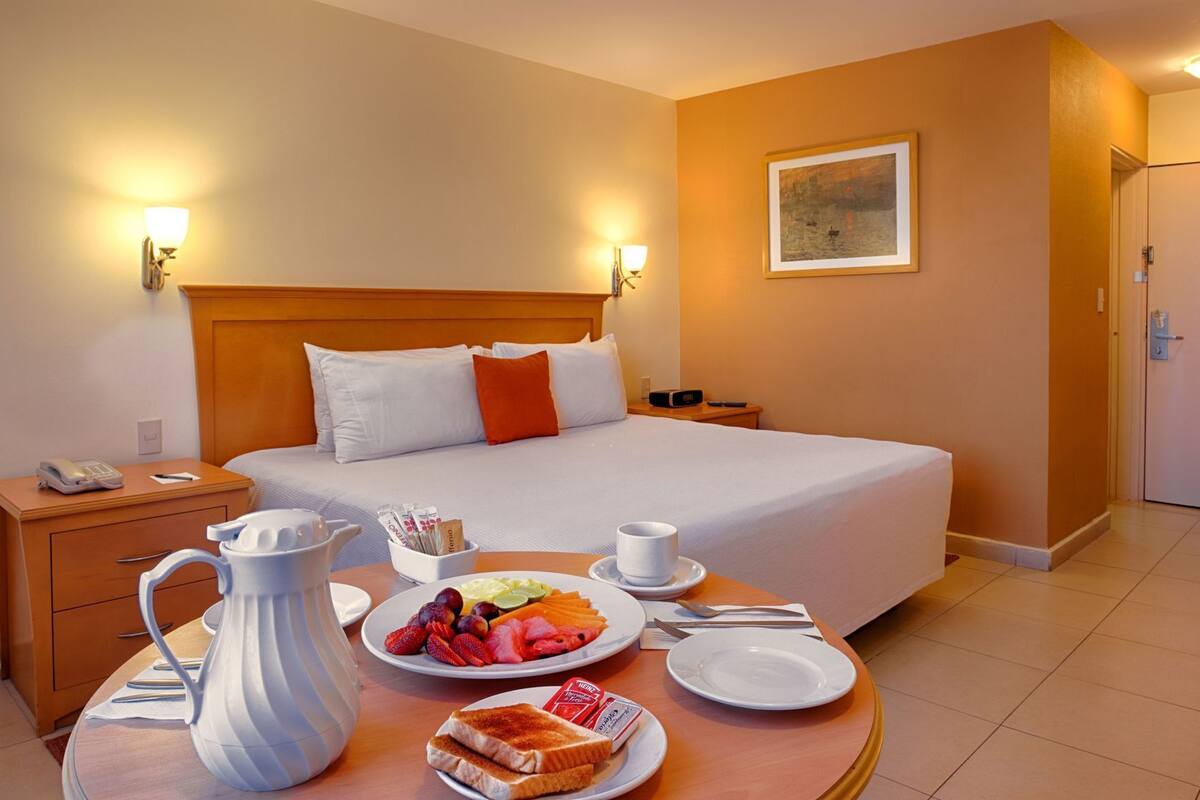 Een hotelkamer met een bed, nachtkastjes, een ontbijttafel met fruit, toast en een karaf, en een schilderij aan de muur.