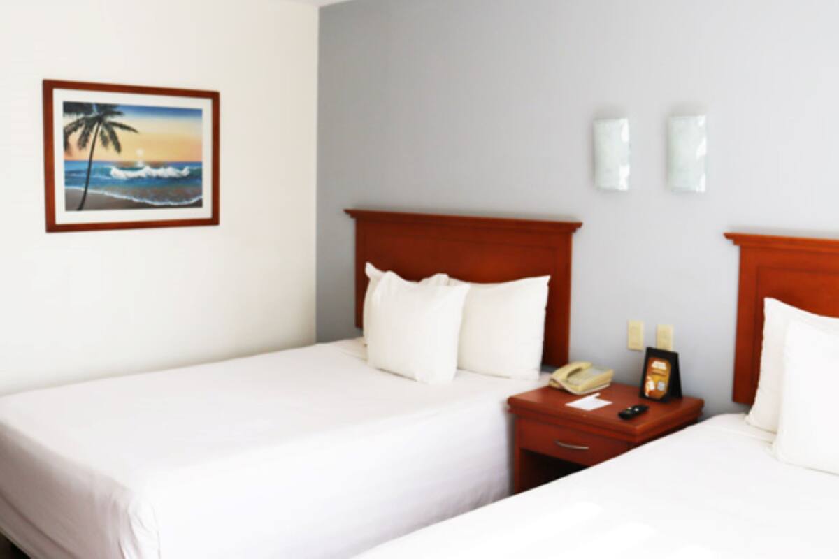 Een hotelkamer met twee eenpersoonsbedden, een houten hoofdbord, een nachtkastje met een telefoon en een klein tafeltje, en een ingelijst strandlandschap aan de muur.