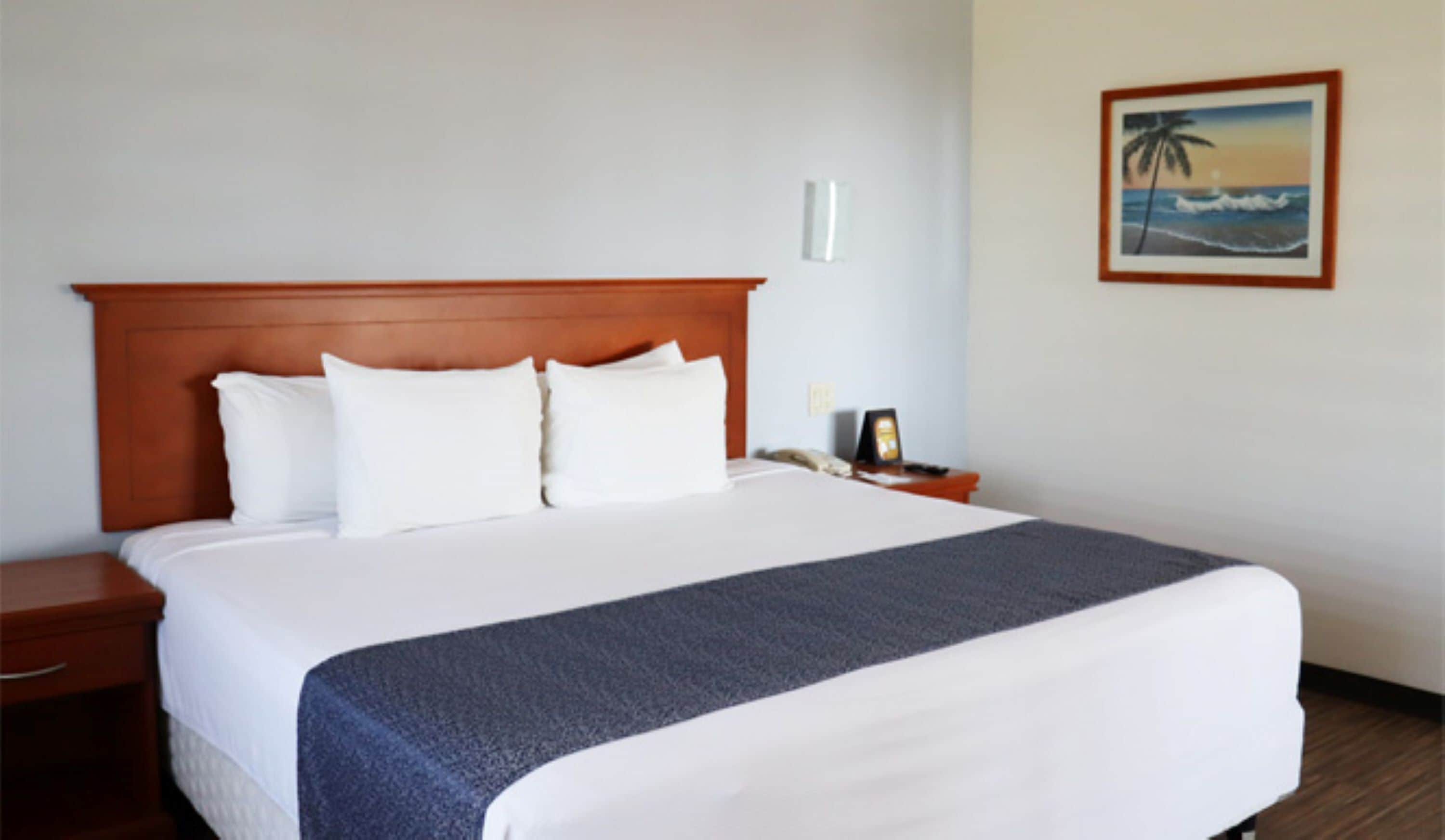 Habitación de hotel con una cama grande, cabecera de madera, ropa de cama blanca, una mesita de noche con teléfono y un cuadro enmarcado con una escena de playa en la pared.