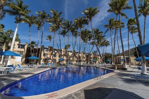 Una piscina rodeada de palmeras y sombrillas azules, con sillas de descanso y un edificio al fondo.