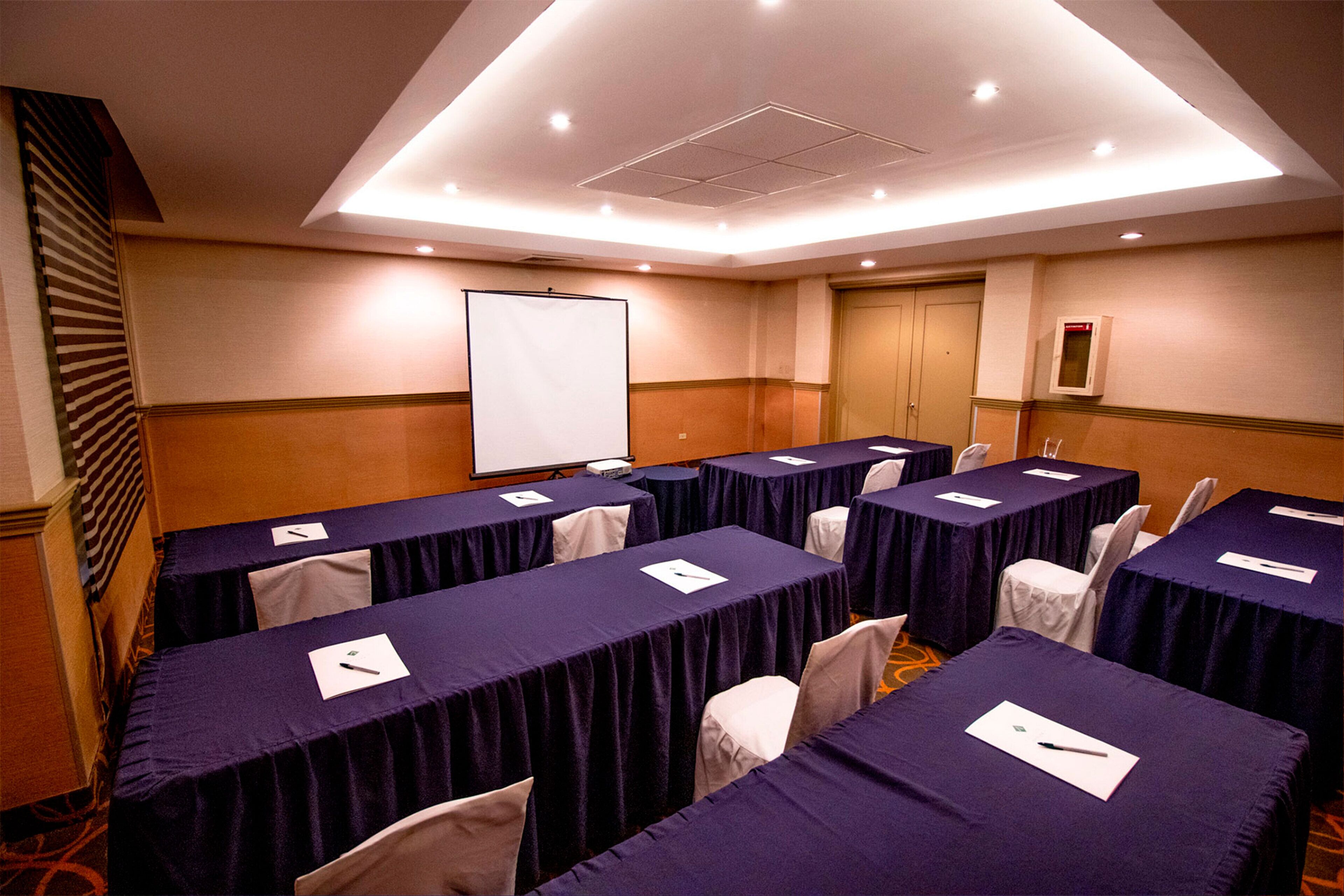 Uma sala de conferências com várias mesas cobertas por toalhas roxas, cadeiras brancas e um quadro branco na parede.