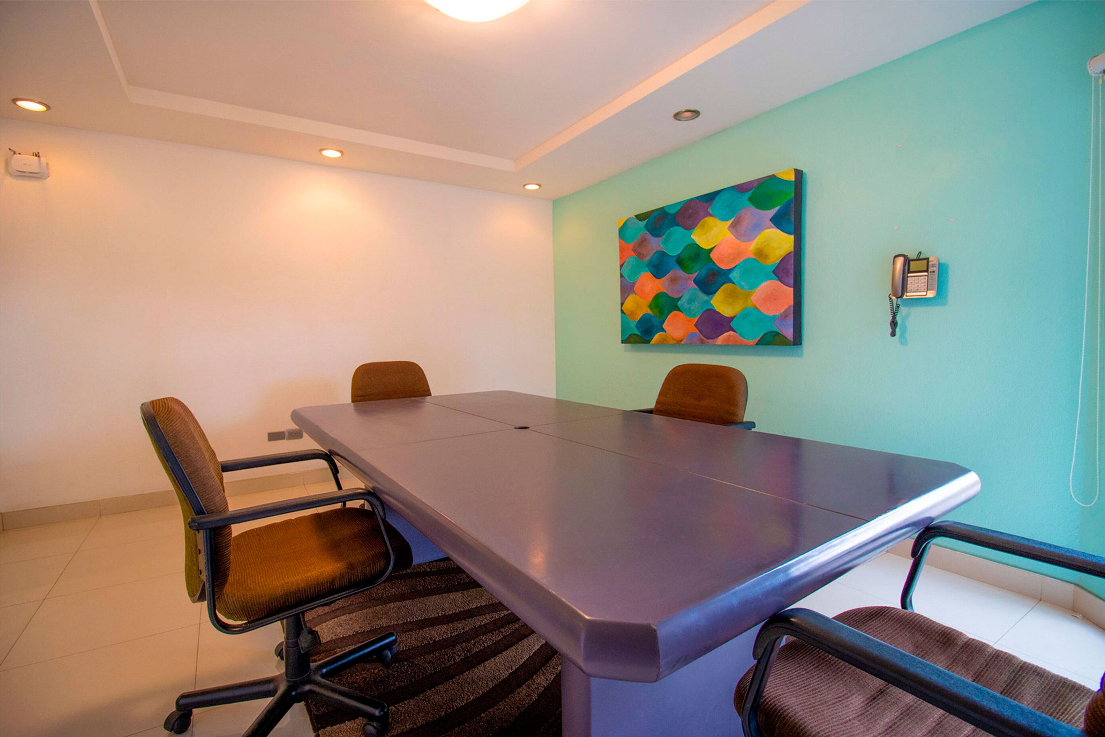 A conference room with a long table, four chairs, a colorful abstract painting on the wall, and a telephone.