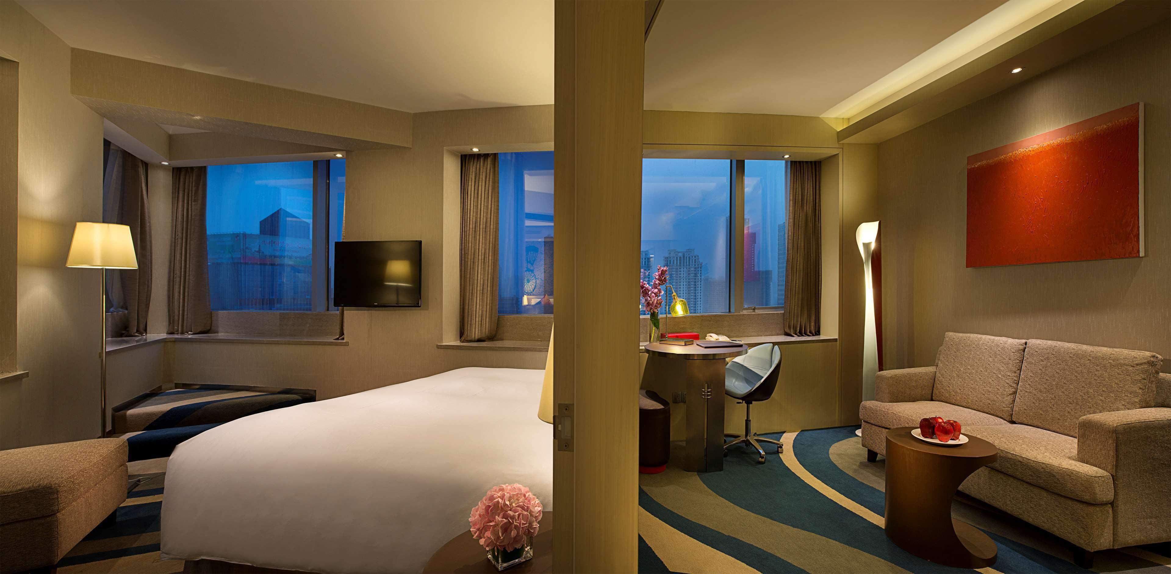 A hotel room with a large bed, a desk with a chair, a sofa, a TV, and a window with a city view.