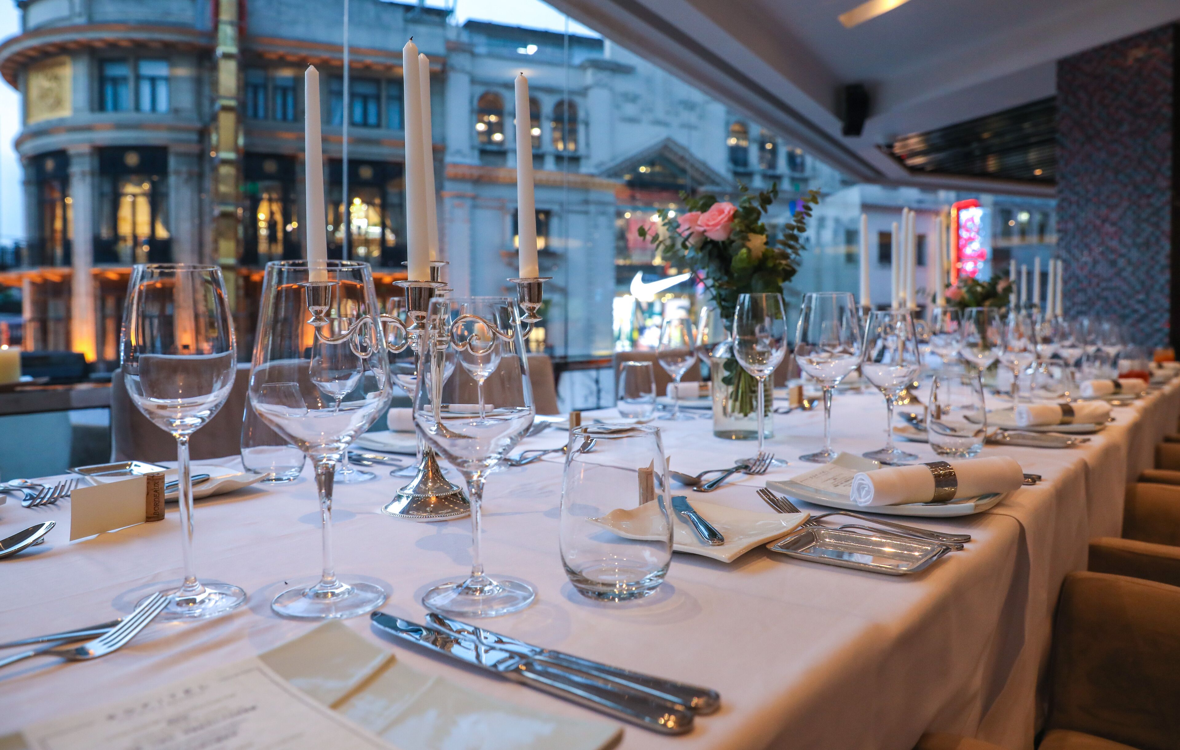 A long dining table set with multiple wine glasses, candles, and cutlery, with a view of a historic building in the background.
