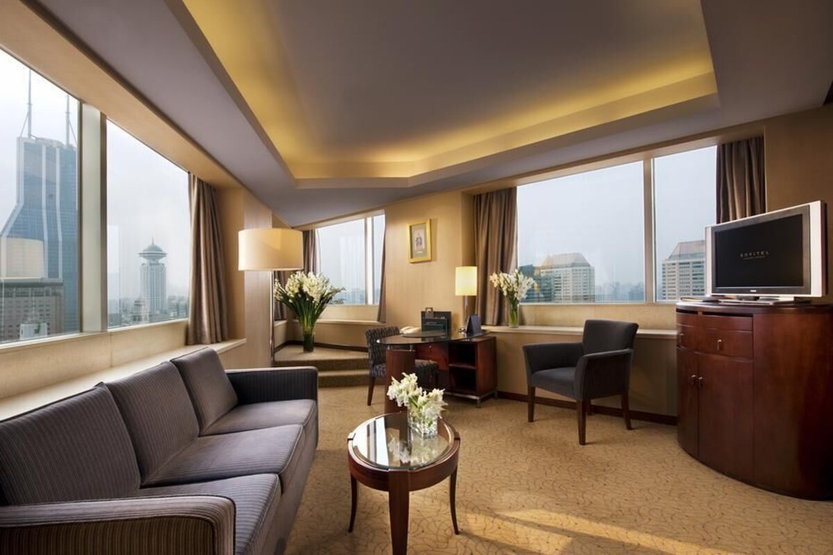 A hotel room with a sofa, armchair, TV, desk, and large windows with a city view.