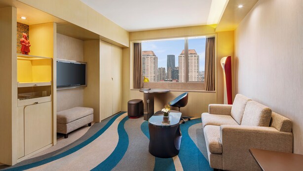 Habitación de hotel con sofá, sillón, televisor y vista a la ciudad.