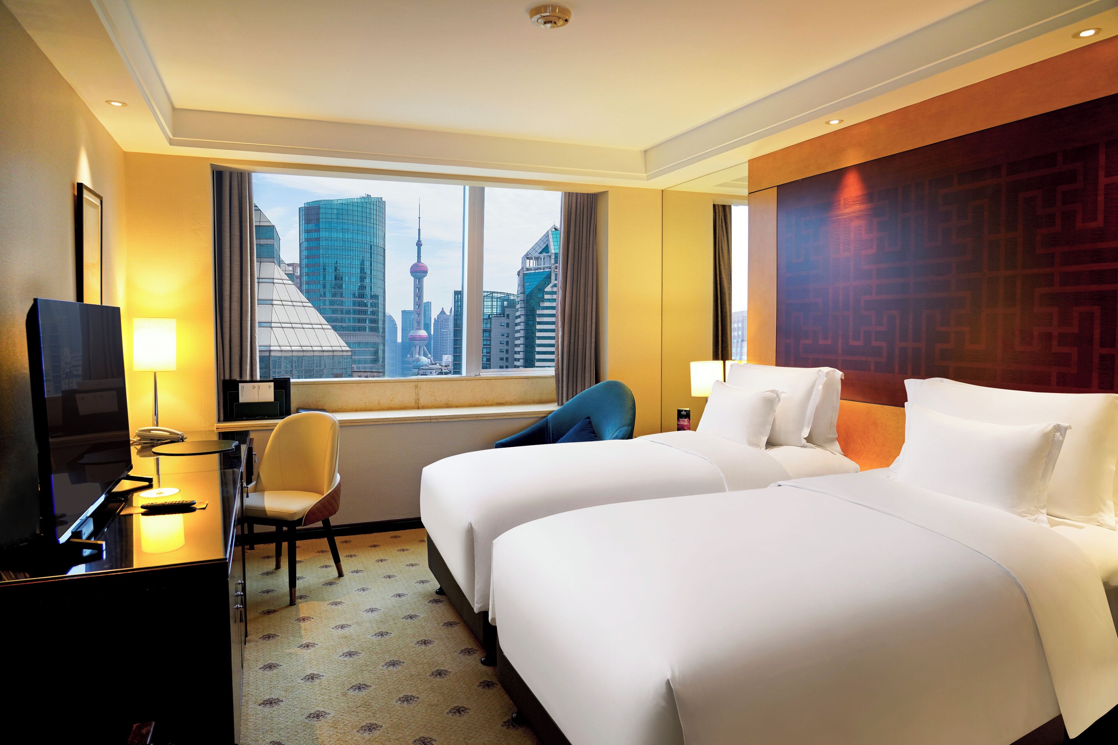 A hotel room with two beds, a desk with a computer, a chair, a TV, and a window with a city view.