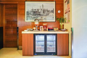 A hotel reception area with a wooden panel wall, a framed cityscape picture, a beverage dispenser, and a refrigerator stocked with bottled water.