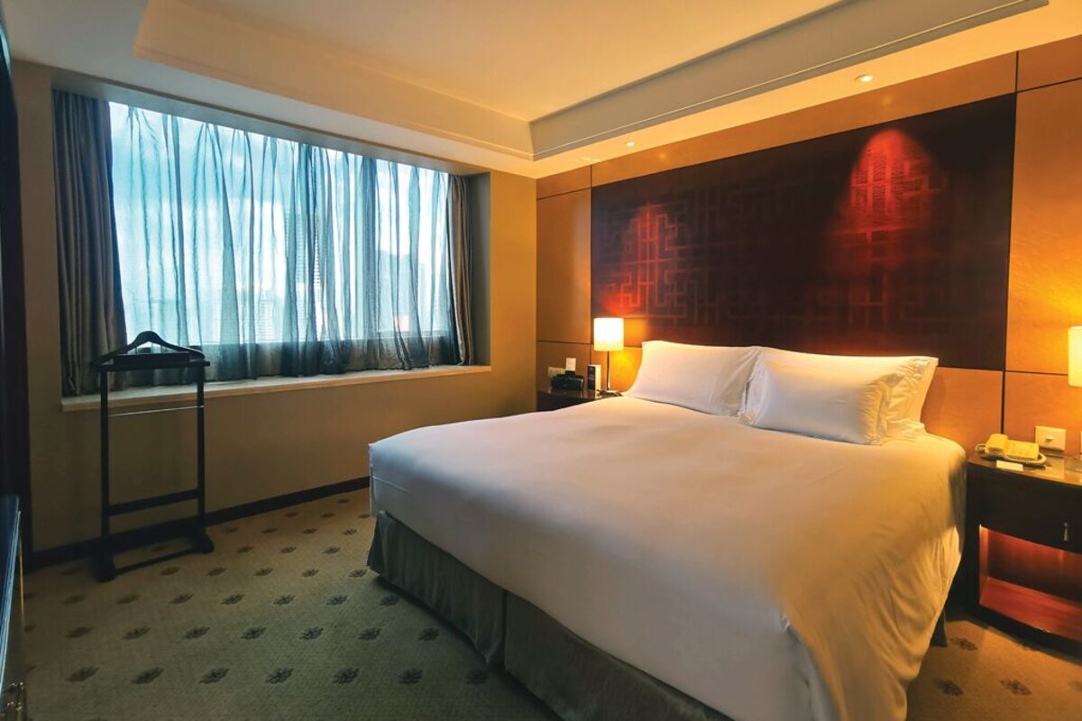 A hotel room with a large bed, a window with sheer curtains, a bedside table with a lamp, and a wall-mounted artwork.