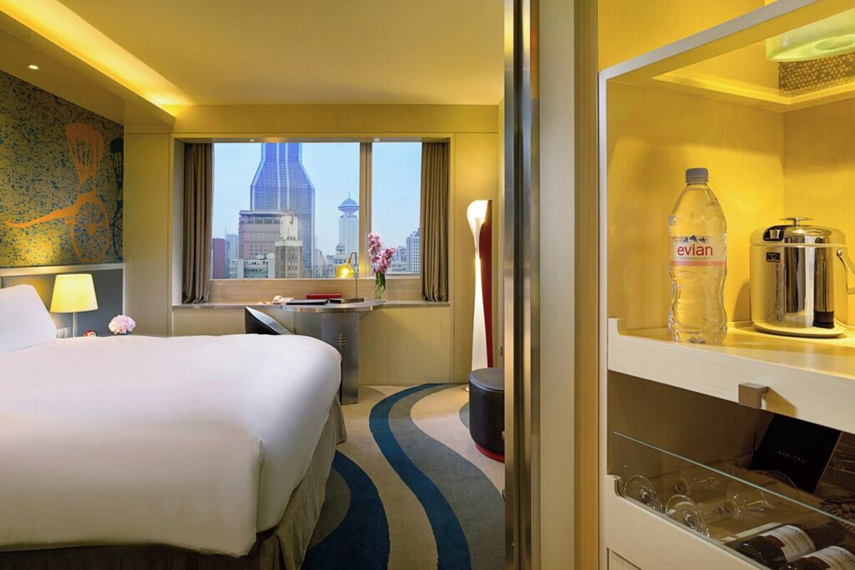 A hotel room with a bed, a desk, a window with a city view, a minibar, and a glass cabinet.