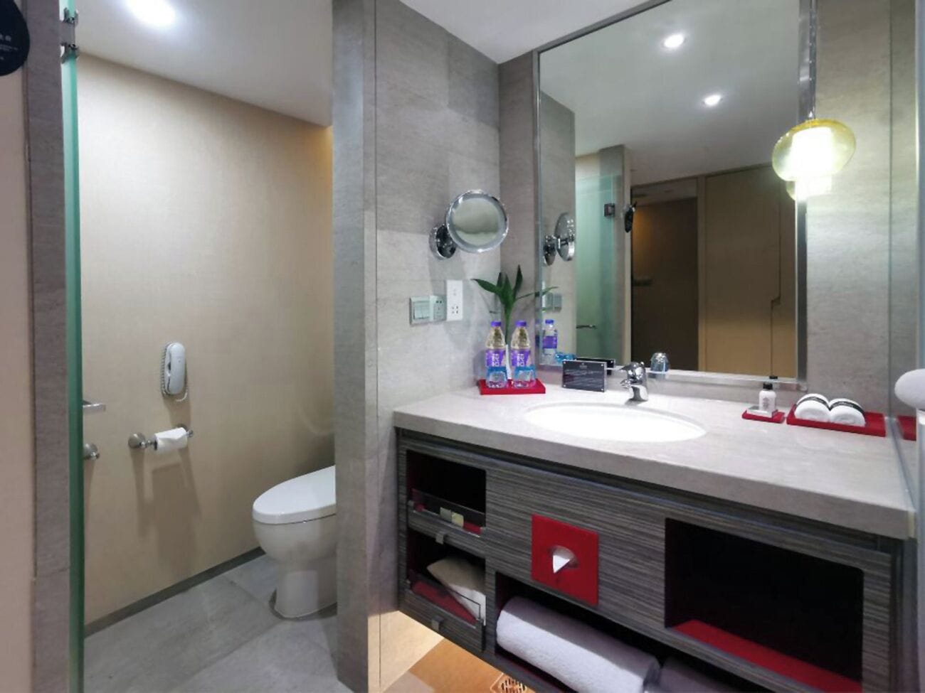 Un baño moderno con inodoro, lavamanos y espejo. Hay artículos de aseo en la encimera y un perchero.