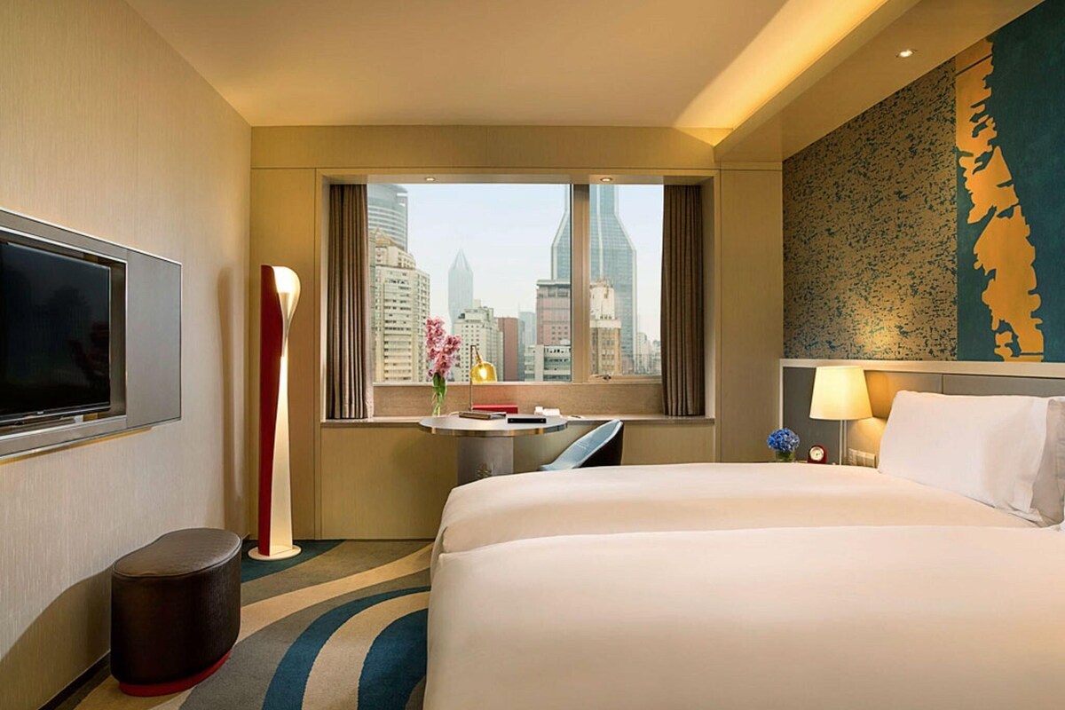 A hotel room with a bed, a desk, a TV, a window with a city view, and a decorative wall.
