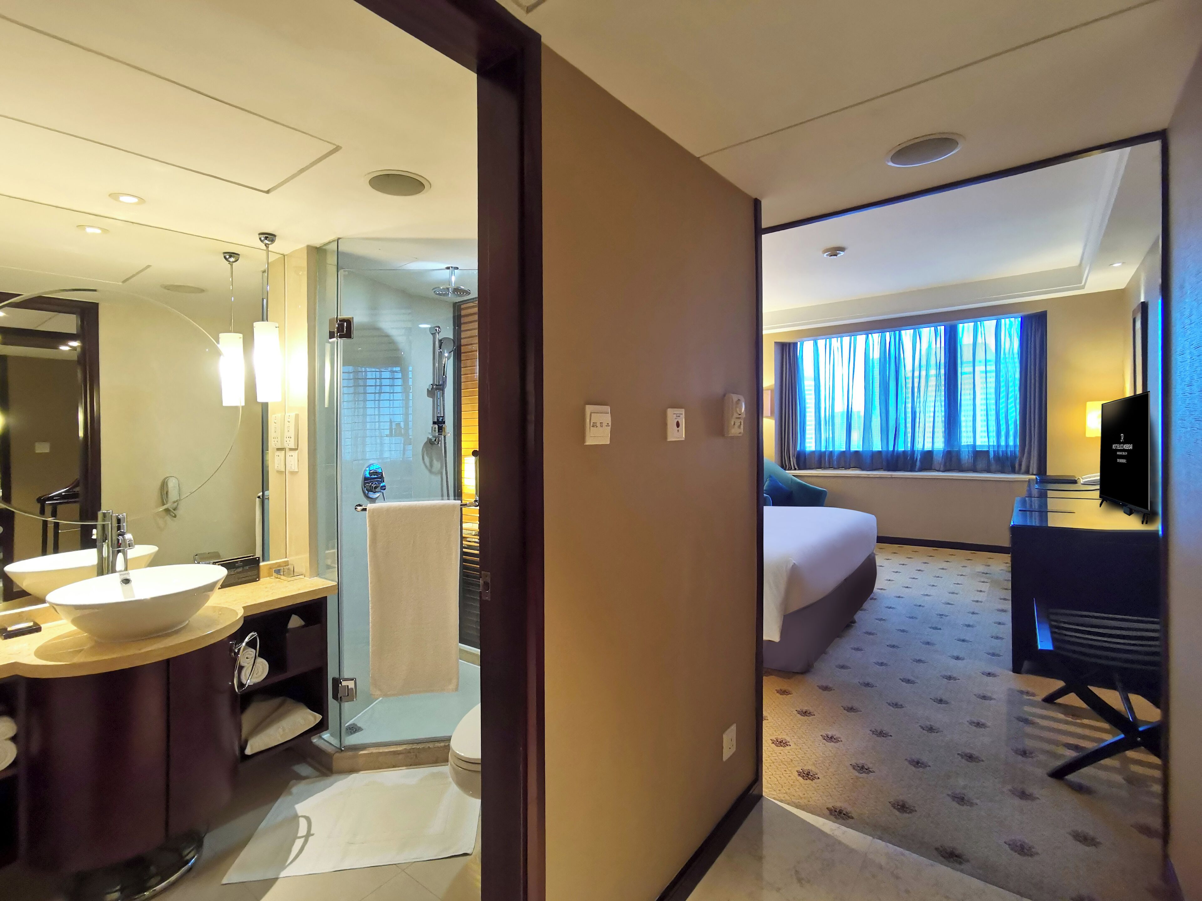 A hotel room with a bathroom, a bed, a desk, and a TV.