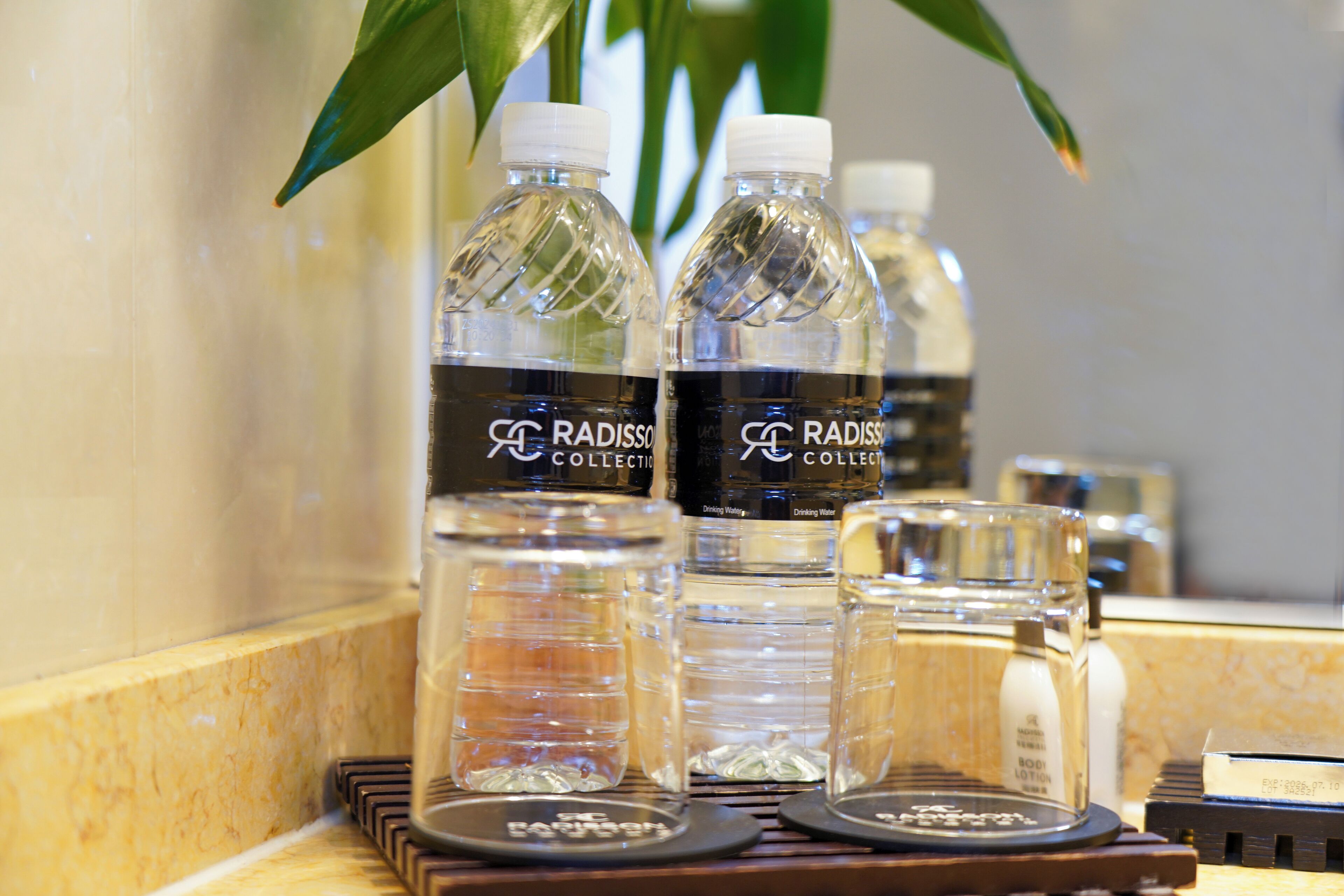 A bathroom counter with three bottles of Radisso Collection drinking water, a small glass, and a bottle of body lotion.