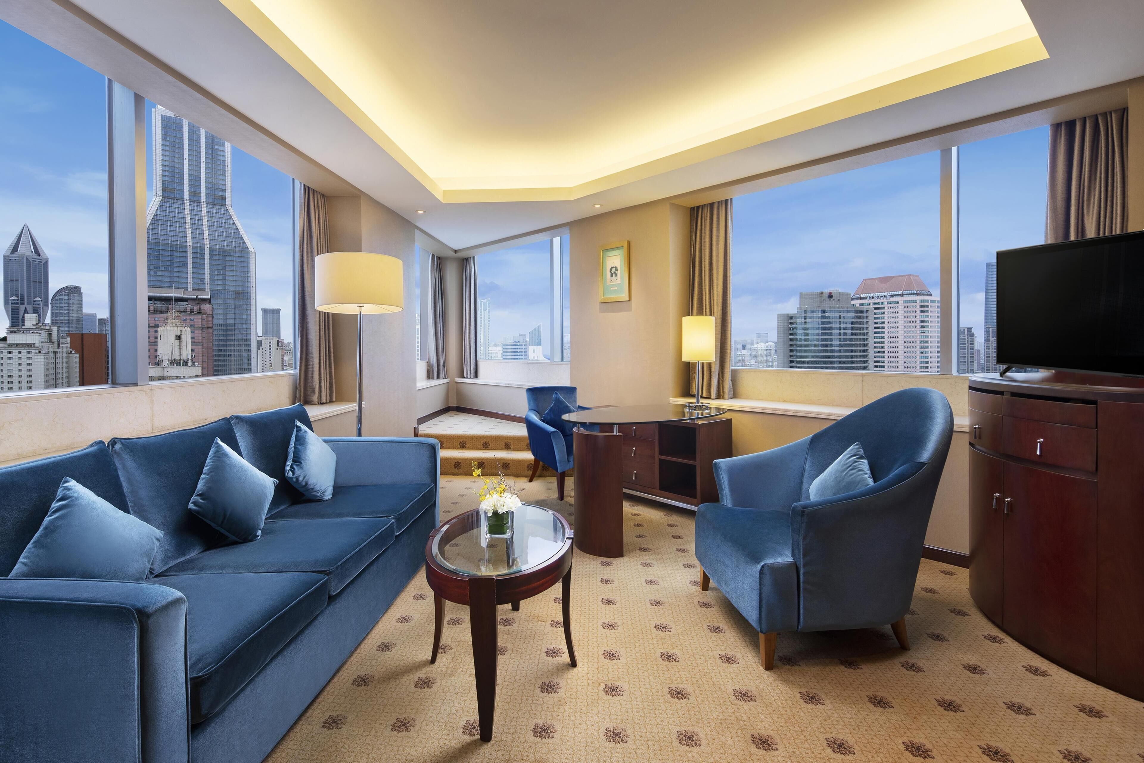 Une chambre d’hôtel équipée d’un canapé, d’un fauteuil, d’un bureau, d’une télévision et de grandes fenêtres donnant sur la ville.