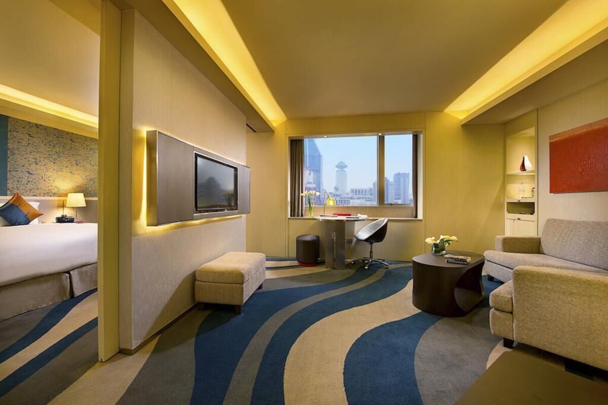 A hotel room with a bed, a desk, a chair, a sofa, a TV, and a window with a city view.