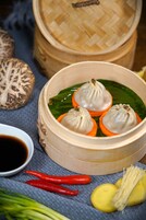 A bamboo steamer with dumplings on a leaf, a bowl of dipping sauce, red chili peppers, and a mushroom.