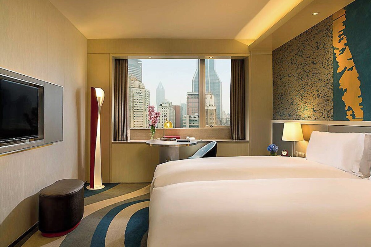 A hotel room with a bed, a TV, a desk, a lamp, and a view of the cityscape.