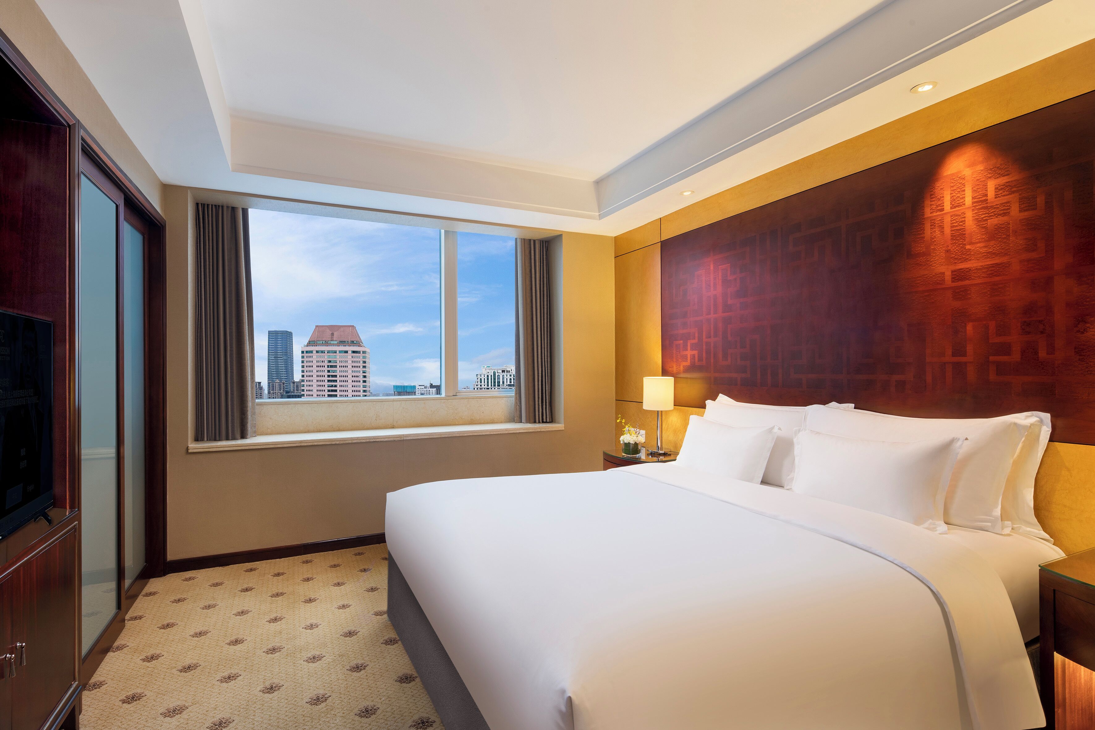 A hotel room with a large bed, a TV, a window with curtains, and a city view.