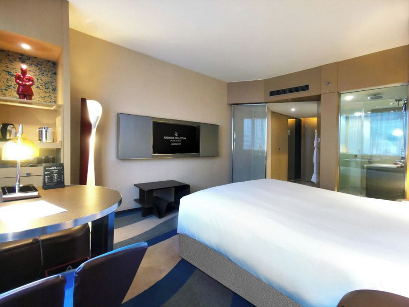 A modern hotel room with a large bed, a desk with a lamp, a flat-screen TV, and a glass shower enclosure.