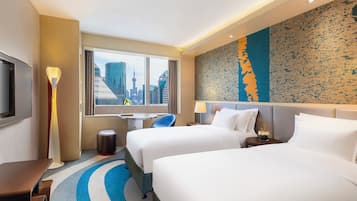 A hotel room with two beds, a desk, a chair, a TV, and a large window with a city view.