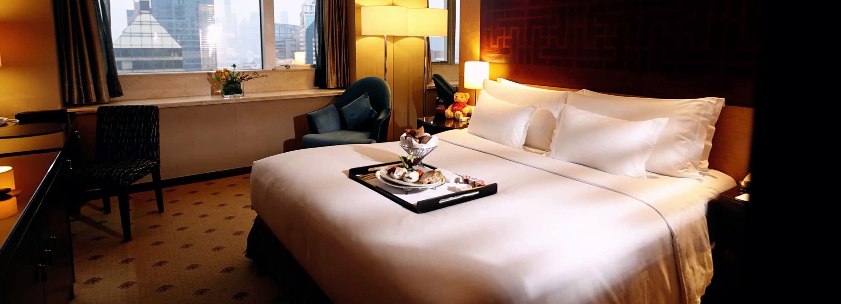 Ein Hotelzimmer mit einem großen Bett, einem Schreibtisch mit Lampe, einem Sessel und einem Fenster mit Blick auf Gebäude.