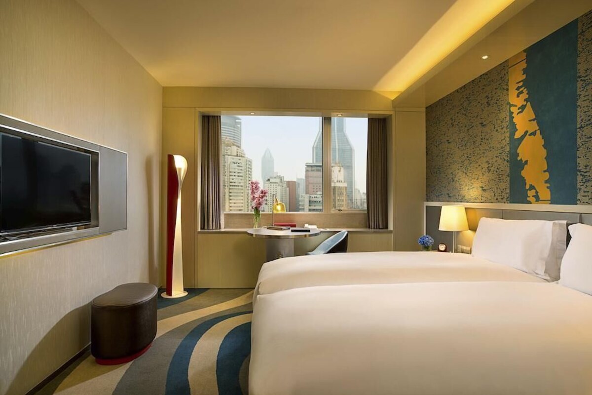 A hotel room with a large bed, a TV, a desk, a lamp, and a view of the city skyline.