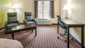 Een hotelkamer met twee fauteuils, een tafeltje, een bureau met stoel, een raam met ruitjesgordijnen en een airconditioning.