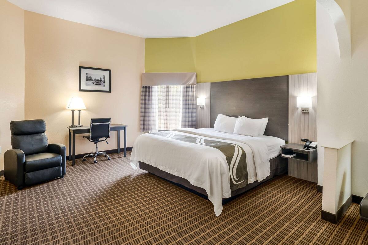 Una habitación de hotel con una cama grande, un escritorio con silla, una lámpara y un cuadro en la pared.