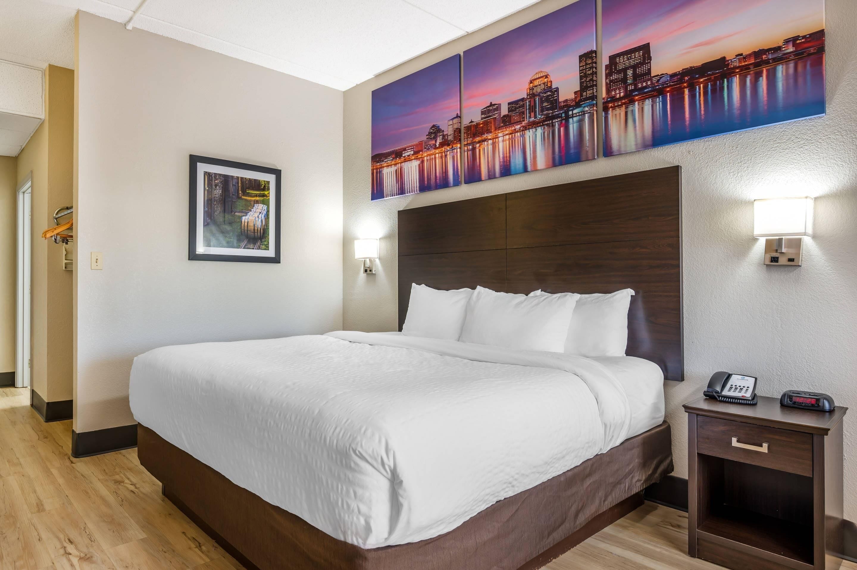 Habitación de hotel con una cama grande, una mesita de noche con un teléfono y un reloj, una lámpara de pared y un cuadro en la pared.