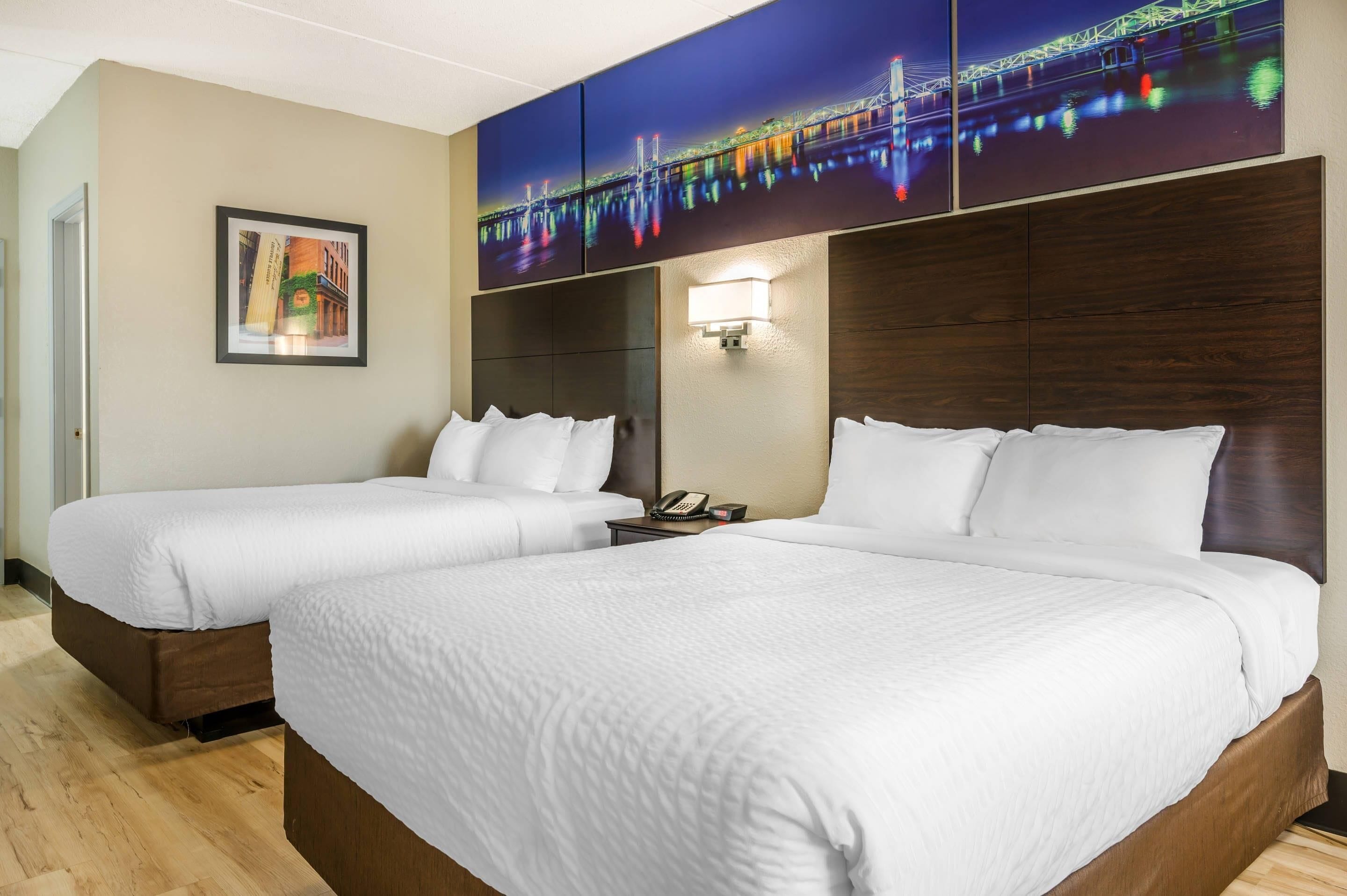 Quarto de hotel com duas camas, mesa de cabeceira, telefone fixado na parede e um quadro emoldurado.