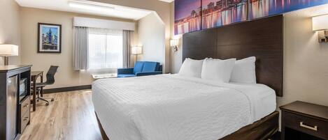 A hotel room with a large bed, a desk with a chair, a blue armchair, and a cityscape painting on the wall.