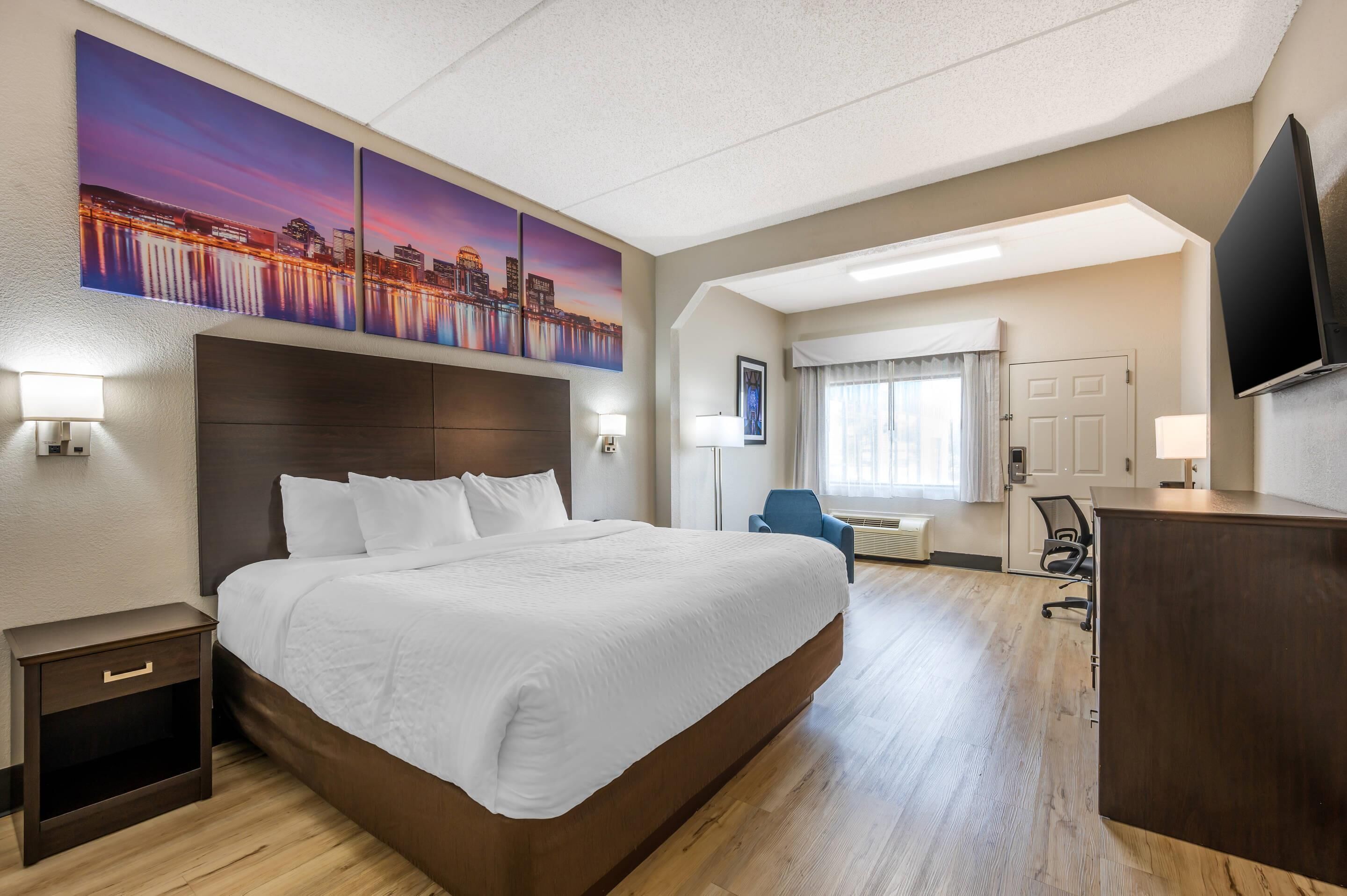 Ein Hotelzimmer mit einem großen Bett, einem Schreibtisch, einem Sessel, einem Fernseher und Bildern an der Wand.