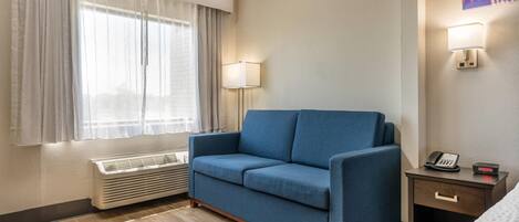 A hotel room with a blue sofa, a bedside table with a phone and a digital clock, a window with curtains, and a wall-mounted lamp.