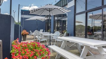 An outdoor patio area with white picnic tables and striped umbrellas, surrounded by a blue building with large windows.