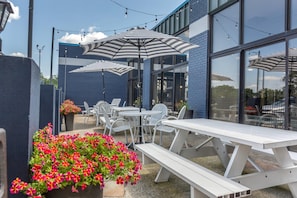 An outdoor patio area with white picnic tables and striped umbrellas, surrounded by a blue building with large windows. - Clarion Pointe Louisville East (Louisville)