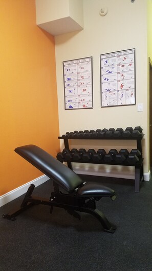 A gym with a bench and dumbbells, two instructional posters on the wall, and a door in the background. - Clarion Pointe Louisville East (Louisville)