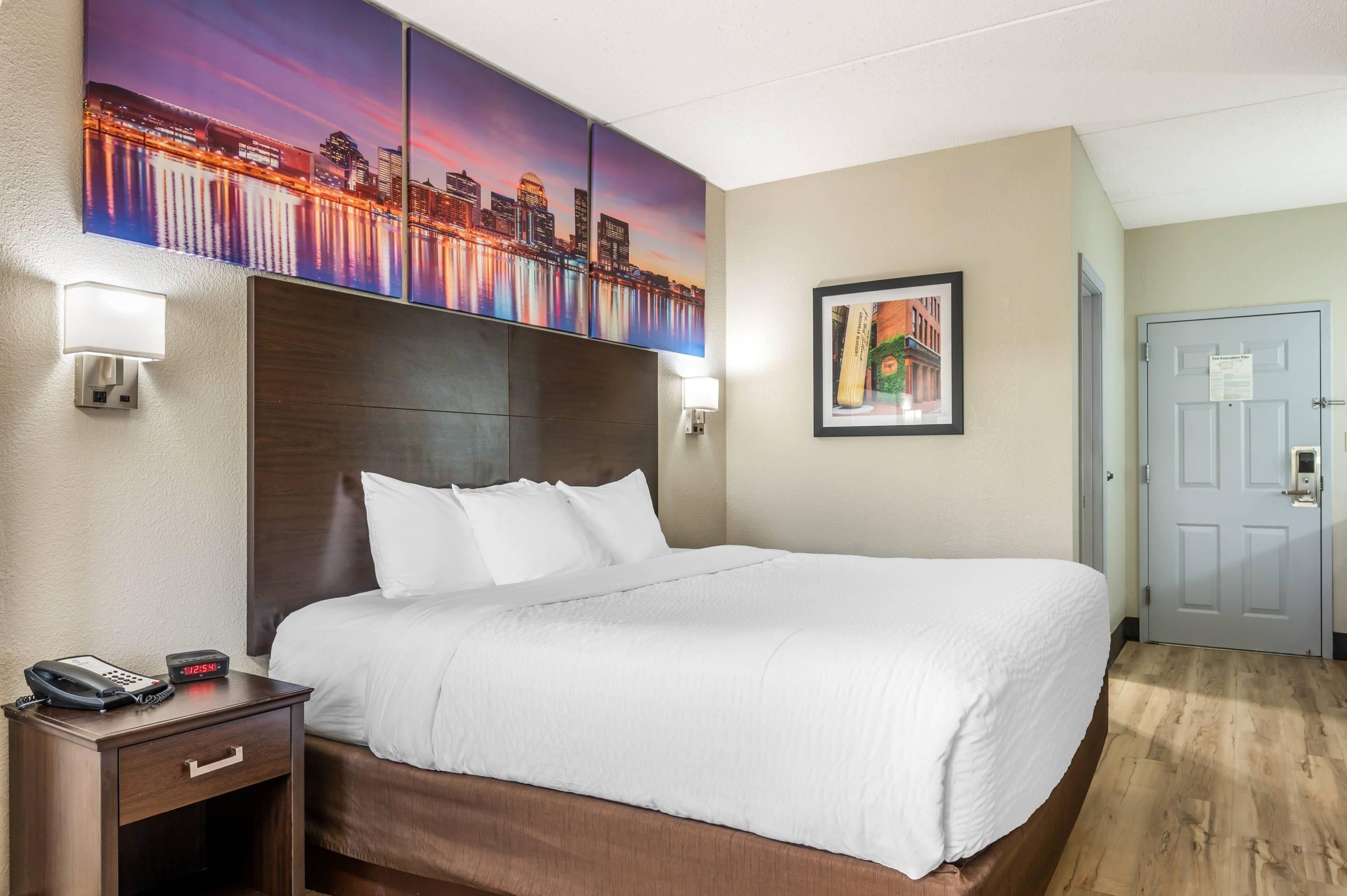 Habitación de hotel con una cama grande, una mesita de noche con un teléfono, un cuadro de un paisaje urbano y una puerta con el plan de evacuación contra incendios.