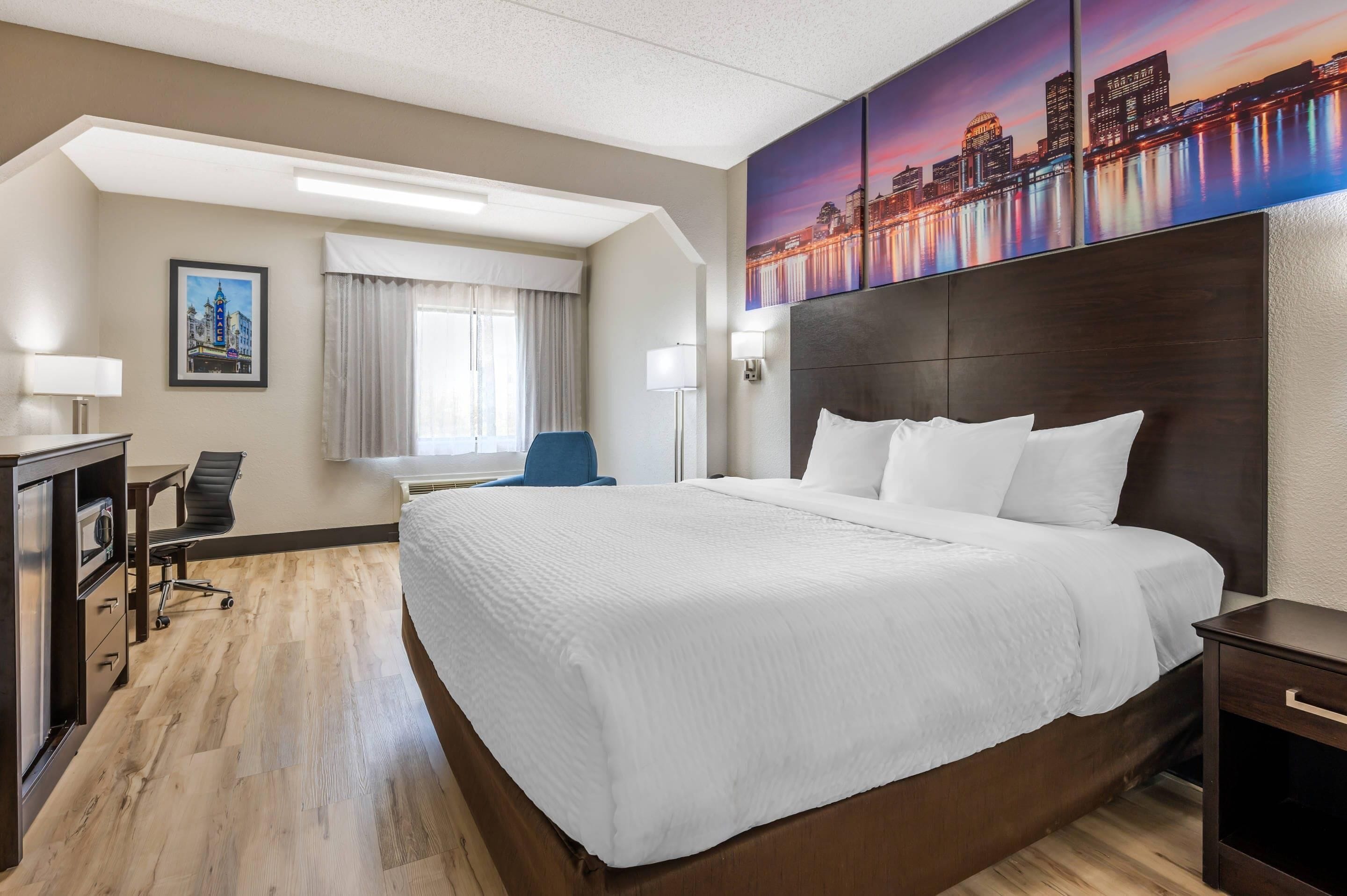 A hotel room with a large bed, a desk with a chair, a window with curtains, a painting of a cityscape, and a nightstand.